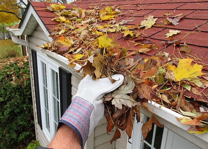 A gloved hand removing leaves from the gutter of a red-roofed house, illustrating the importance of roof maintenance by General Roofing Company in Oakland.